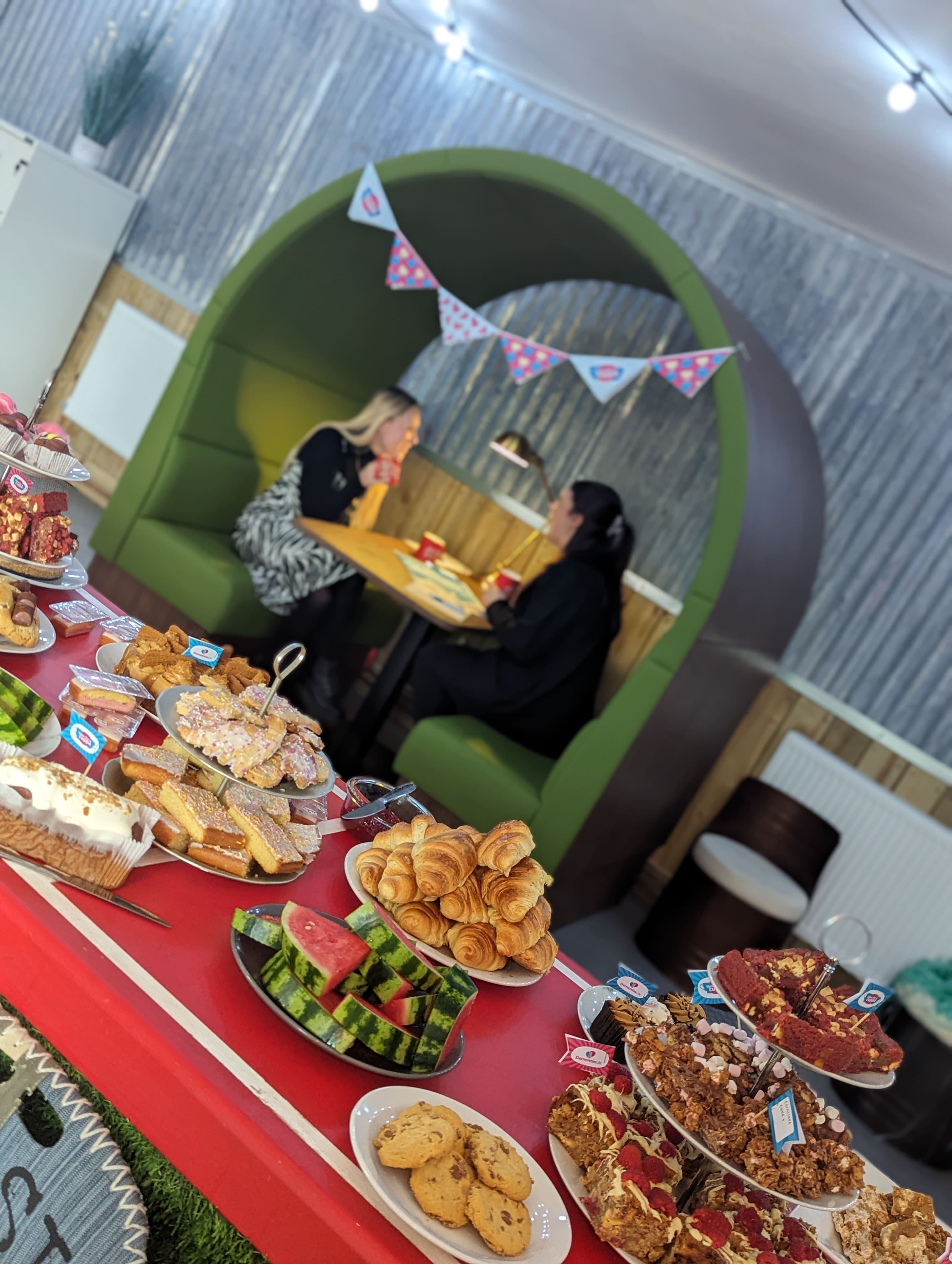 2 women chatting in the background with a spread of cakes in the foreground.