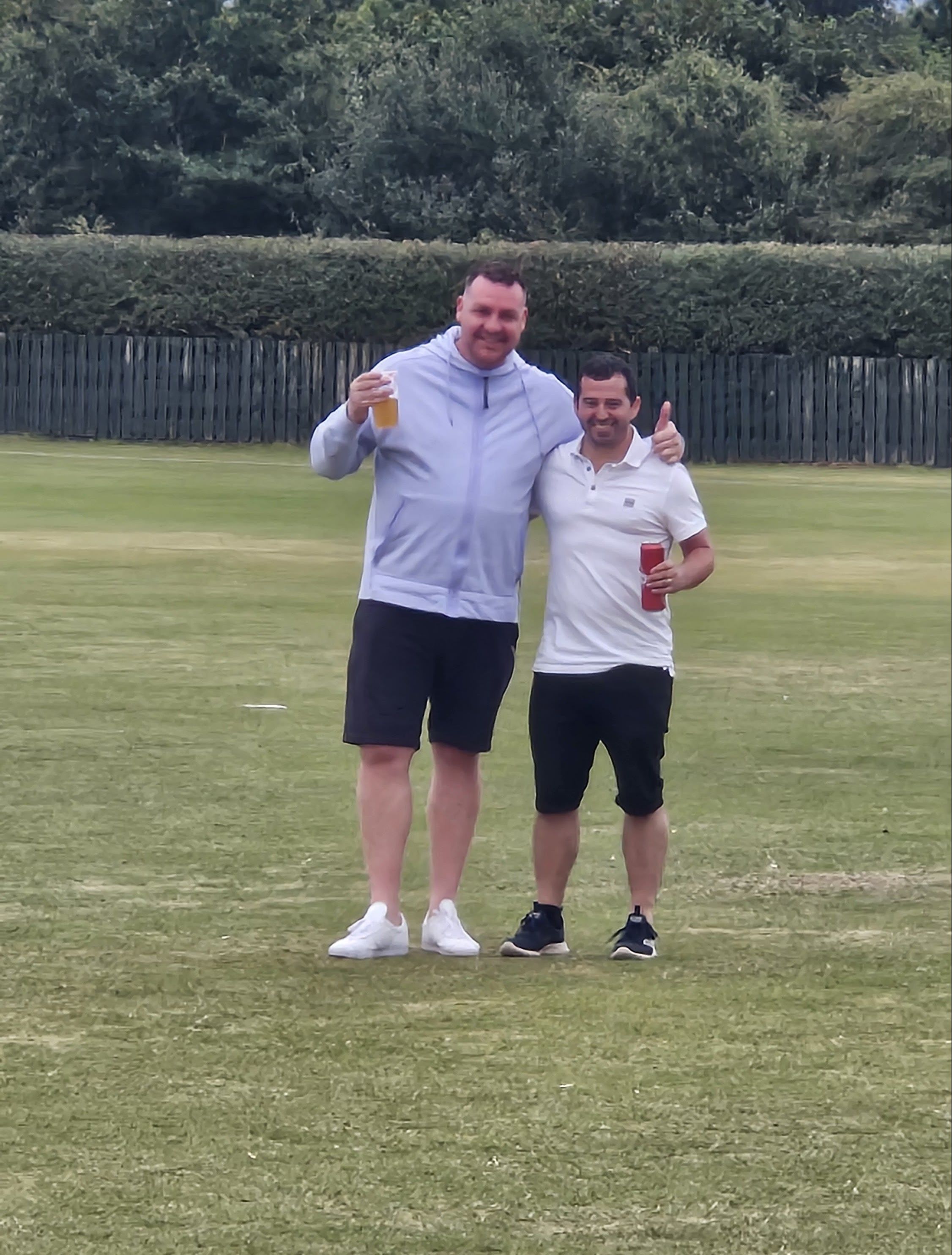 2 men on a cricket field holding drinks and smiling.